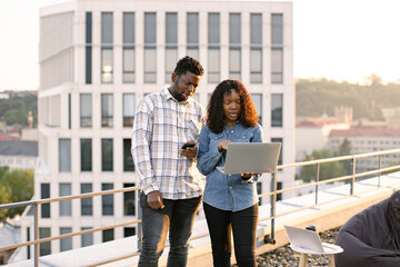 Two African American business people outdoors during break standing on rooftop. Young adult man and woman using laptop while standing outdoors office building.