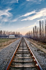 Fototapeta premium A railroad disappearing into the distance amidst fields in rural scenery, accompanied by old wooden electrical poles running alongside, against the backdrop of a sunset sky and distant light mist.