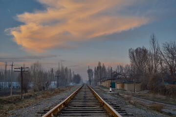 Fototapeta premium A railroad disappearing into the distance amidst fields in rural scenery, accompanied by old wooden electrical poles running alongside, against the backdrop of a sunset sky and distant light mist.