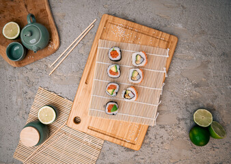 Salmon sushi rolls on bamboo sushi mat and wooden cutting board, chopsticks, fresh lime, green teacup on a modern concrete countertop. Restaurant branding, or culinary content creation.