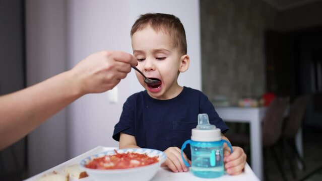 Adorable baby opens his mouth wide waiting for the spoon from his mom. Toddler is given borsch for lunch. Blurred backdrop.
