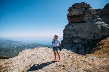 A female tourist stands on a cliff on a cliff against the background of a mountain landscape. The girl raised her head to the sky. She's wearing a white T-shirt and denim shorts