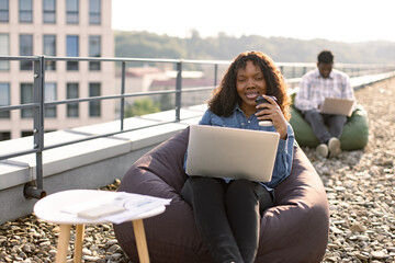 Charming African American lady in casual shirt holding cup of hot drink while scrolling webpages on portable computer outdoors, while male colleague working in background.