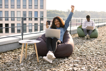 Delighted African American woman screaming and raising hand up looking at laptop during coffee break on rooftop while her colleague working in blurred background.