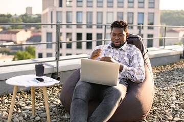 Obraz premium Happy African American person in casual clothing gesture at laptop camera on flat roof of building. Efficient freelancer beginning video conference via internet connection outdoors.