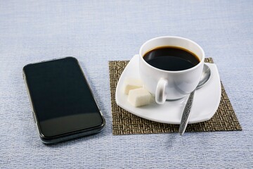 A white cup of black coffee with sugar on a napkin-mat and a phone on the table early in the morning.