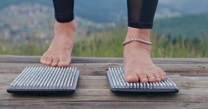 Unrecognizable female yogi standing on sadhu boards with sharp nails outdoors. Close up of female in black leggings, massaging feet, using sadhu boards, in slow motion. Alternative medicine concept. 