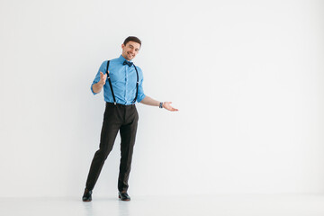 A man in a blue shirt, classic trousers, suspenders and a bow tie against a white wall in the...