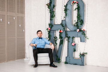 A man in a blue shirt, classic trousers, suspenders and a bow tie on the background of a huge...