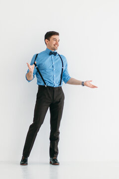 A man in a blue shirt, classic trousers, suspenders and a bow tie against a white wall in the studio. The man pulls off the suspenders with one hand