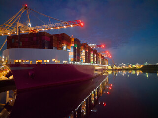 Fototapeta na wymiar Pink container ship at night at Southampton Docks, UK. Aerial view of the bow and side view of the vessel loaded with shipping containers. Calm sea with ship and port crane lights reflection.