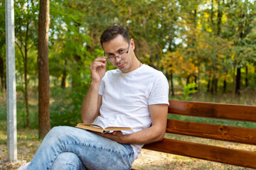 A guy in glasses sits on a bench in the park and reads a book