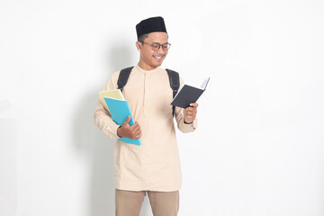 Portrait of excited student Asian muslim man in koko shirt with skullcap carrying school books, while reading his notebook. Isolated image on white background