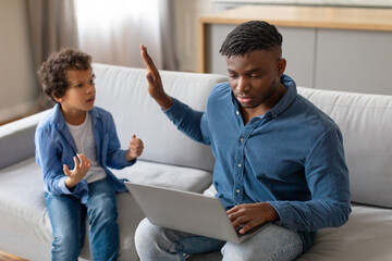 Black boy gesturing to distracted father on laptop