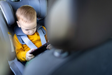 Cute boy watching cartoon or playing game on mobile phone while sitting in the car.
