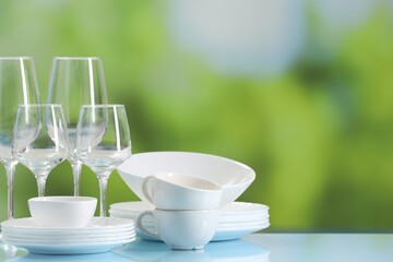 Set of many clean dishware and glasses on light blue table against blurred green background. Space for text