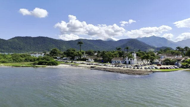 Aerial shot drone flies to the right over water with a white church on the beach and a jetski that zooms through the frame