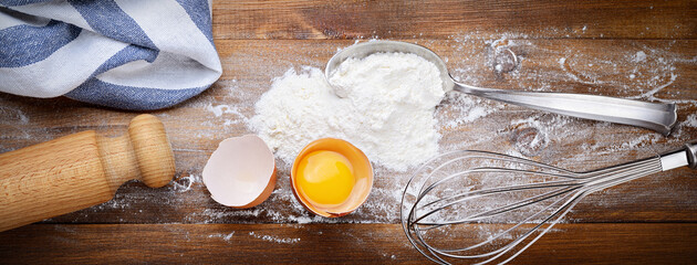 Egg and flour with spoon, stainless steel whisk and rolling pin on wooden background, top view.