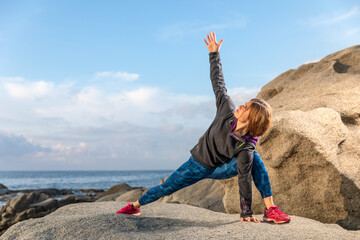 Young woman dressed in sports clohing, in a extended side angle position, on some rocks with the sea in the background