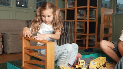 Caucasian girl sits at sand box while placed car model at slope toy. Diverse children playing plastic model while sitting at school in break time at play room. Creative activity concept. Erudition.