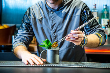 Barman prepara un mojito cocktail en la barra de un cocktail bar exclusivo vestido con una chaquetilla azul con iluminación nocturna, detalle close up