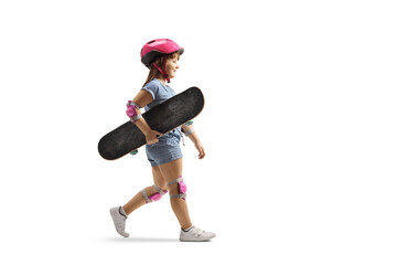Full length profile shot of a little girl walking and carrying a skateboard, wearing elbow, knee pads and helmet