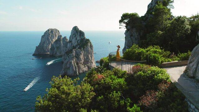 Iconic Faraglioni rocks viewed from a hiking trail on Capri. A lone observer woman takes in the splendor of the towering sea stacks, picturesque seascape.