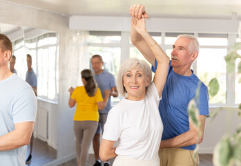 Fototapeta na wymiar Positive senior man dancing playful Latin dance bachata with female partner during group class in choreography studio. Active lifestyle of older generation