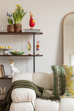 Sunny and design interior of living room with easter decorations, tulips, modular sofa, shelf, pillows and elegant accessories. Home decor. Easter holidays. 