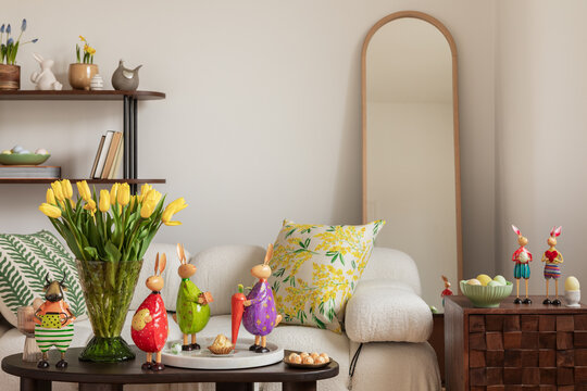 Sunny and design interior of living room with easter decorations, tulips, modular sofa, shelf, pillows and elegant accessories. Home decor. Easter holidays. 