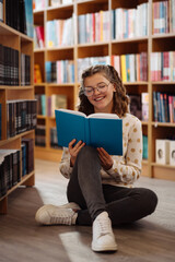 Smiling teenage girl in glasses enjoys reading a blue book, seated on the floor in a vibrant library