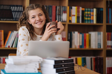 A radiant teenage girl holding a mug, enjoys a moment's break from studying on her laptop in the library