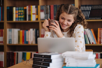 Teenage girl beams with a smile, holding a mug while engaging with her laptop in a library full of...