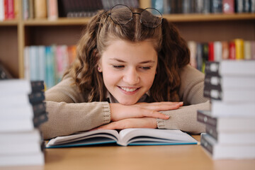 A teenage girl with glasses perched on her head is captivated by a book in the library, with a joyful expression