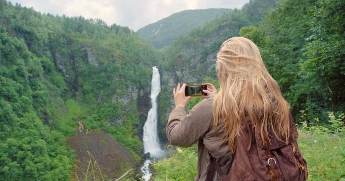 Photography, tourist or woman taking pictures in nature for travel, adventure or sightseeing scenery. Waterfall, lady or photographer taking photo for outdoor destination memory or location in Norway