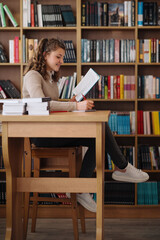 A teenage girl with a smile sits at a library table, absorbed in her book with notes and a laptop...