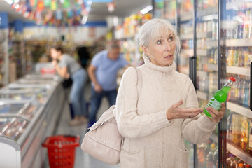 Positive casual aged woman in knitted dress walking down aisle of grocery store, choosing refreshing non-alcoholic beverages