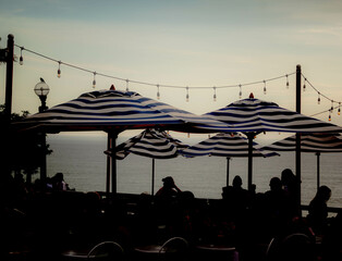 Sunset Silhouettes: A captivating scene at a restaurant as the sun sets, casting a warm glow on patrons beneath umbrellas. The silhouette of people enjoying a meal against the fading sunlight creates  - obrazy, fototapety, plakaty