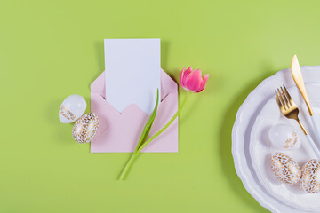 Pink tulips and table place setting with empty greeting or invitation card and envelope on pastel green background. Happy easter card. Top view, flat lay, copy space for text
