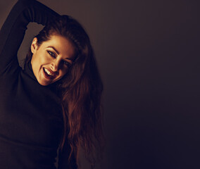 Enjoying toothy smiling makeup woman with brown long hair posing in black sweater on dark shadow background with empty copy space. Closeup