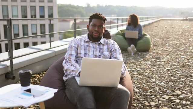 Attractive African American male freelancer using portable computer for remote work at roof top of office, during break sitting in bag chair. Blurred background of businesswoman colleague.