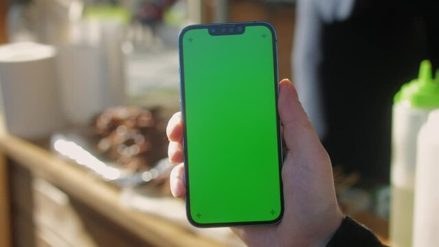 Young man holding phone chroma key screen on the street in front of food market close-up. Male with green screen on smart phone swiping . 