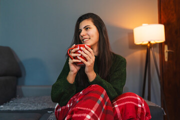 A young girl or woman getting up from bed drinking coffee or tea in blanket cozy room	