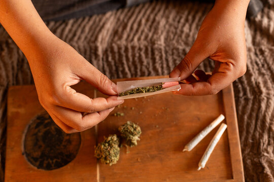 The expert hands of an individual are captured meticulously rolling a joint on a wooden board, with cannabis buds and rolled papers scattered about. The warm, ambient lighting suggests an end-of-day r