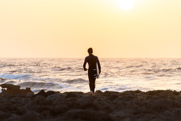 Fototapeta na wymiar Rear View of Young Surfer going to the water Against a Orange Sunset Sky in Canary Island.Copy Space