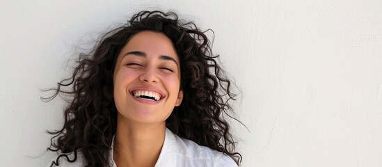A happy woman with dark curly hair smiling, wearing a white shirt, posing in front of a white wall with space for text.