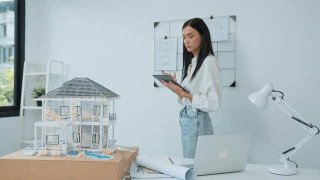 Beautiful caucasian architect focus on using tablet to check house model on meeting table with blueprint, house model and architectural document scatter around at meeting room. Immaculate.