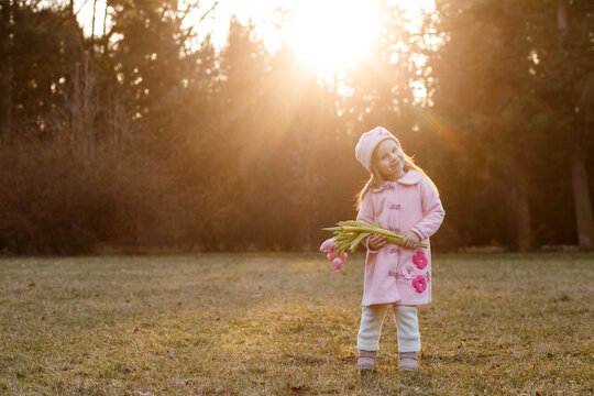 girl 5 years old in a pink coat and hat in retro style with a bouquet of pink tulips in early spring in the park in backlit sunlight.
