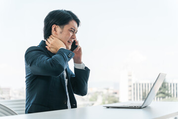 オフィスでノートパソコンを使いながら電話する若いアジア人ビジネスマン（トラブル・困る・クレーム対応）

