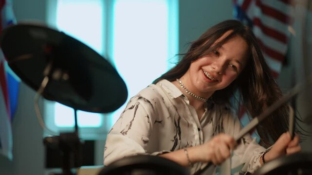 Slow motion. A young girl with braces on her teeth, laughs happily and vigorously taps her drumsticks on an electronic drum machine, while sitting indoors against the backdrop of the American flag